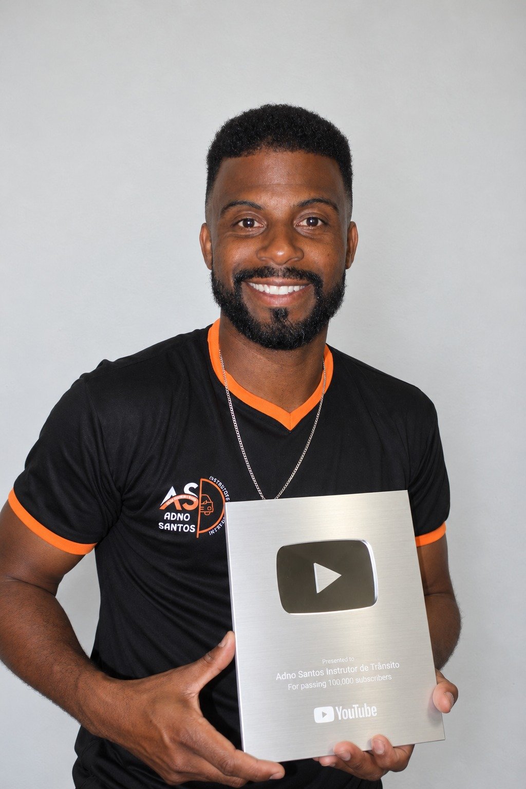Instrutor Adno Santos com uniforme preto e laranja, segurando a placa do YouTube pelo marco de 100 mil inscritos.
